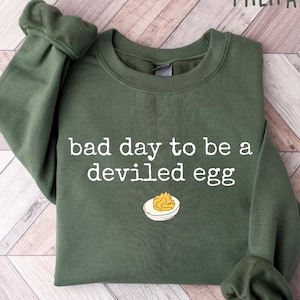 May include: Military green sweatshirt with the text "bad day to be a deviled egg" in white. The design includes a cartoon deviled egg on a plate. The sweatshirt is folded on a light wood surface.