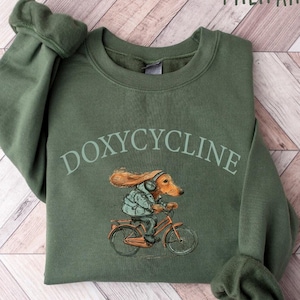 K&ouml;nnte beinhalten: Ein olivgr&uuml;nes Sweatshirt mit dem Aufdruck "DOXYCYCLINE" auf der Vorderseite. Ein Cartoon-Dackel mit Kopfh&ouml;rern f&auml;hrt Fahrrad.
