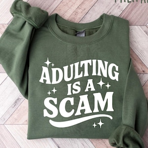 May include: Military green sweatshirt with the text "Adulting is a Scam" in white. The crew neck sweatshirt is folded on a light brown and white wooden surface. The sleeves are cuffed.