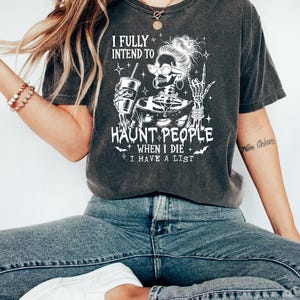 May include: Dark gray t-shirt with the text "I FULLY INTEND TO HAUNT PEOPLE WHEN I DIE I HAVE A LIST" and a skeleton graphic. The skeleton is wearing glasses and holding a drink. The shirt is paired with blue jeans and white shoes.