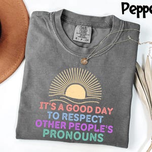 May include: A gray Comfort Colors t-shirt with the text "IT'S A GOOD DAY TO RESPECT OTHER PEOPLE'S PRONOUNS" in colorful letters. The shirt features a sun graphic and a silver necklace. The shirt is a size small.
