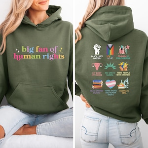May include: An olive green hoodie with the text "big fan of human rights" in colourful letters on the front. The back of the hoodie features various social justice symbols and phrases, including "Black Lives Matter" and "Feminism is for Everyone."