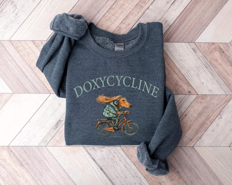 K&ouml;nnte beinhalten: Ein dunkelgrauer Pullover mit dem Text "DOXYCYCLINE" in Gr&uuml;n bedruckt. Auf der Vorderseite des Pullovers ist eine Comic-Illustration eines Hundes auf einem Fahrrad gedruckt.