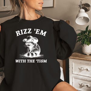 K&ouml;nnte beinhalten: Schwarzer Pullover mit wei&szlig;em Text, der "Rizz 'Em With The 'Tism" lautet, und einem Cartoon-Waschb&auml;ren, der einen Cowboyhut tr&auml;gt.