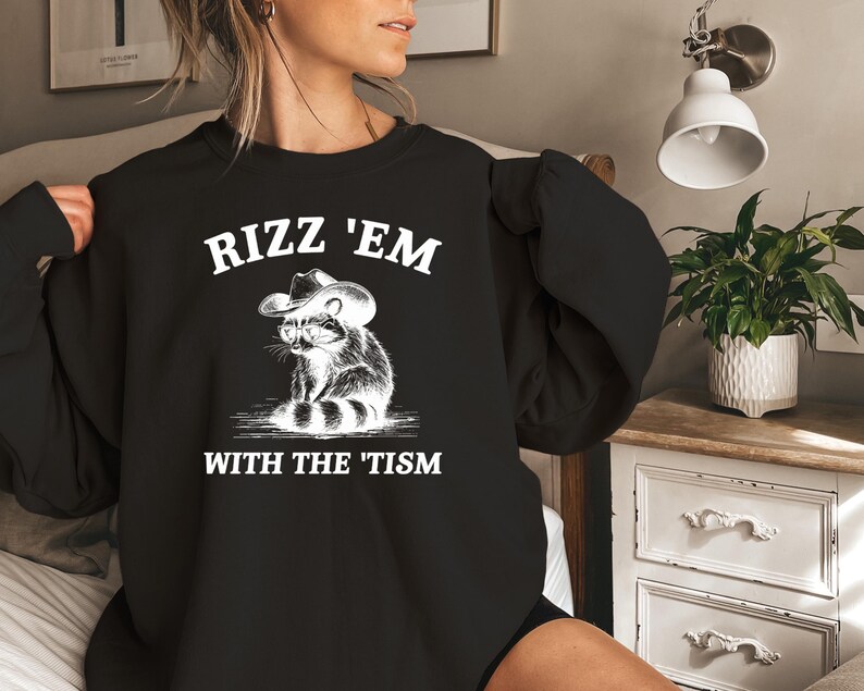 K&ouml;nnte beinhalten: Schwarzer Pullover mit wei&szlig;em Text, der "Rizz 'Em With The 'Tism" lautet, und einem Cartoon-Waschb&auml;ren, der einen Cowboyhut tr&auml;gt.