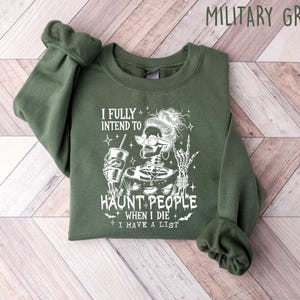 May include: Military green sweatshirt with the text "I FULLY INTEND TO HAUNT PEOPLE WHEN I DIE I HAVE A LIST" and a skeleton graphic. The skeleton is wearing glasses and holding a drink. The sleeves are cuffed.