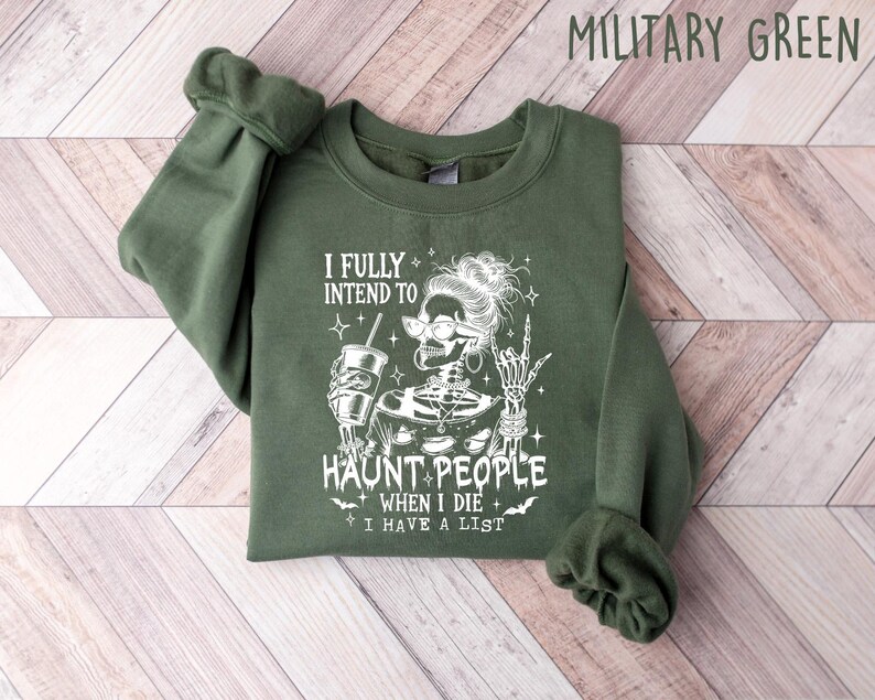 May include: Military green sweatshirt with the text "I FULLY INTEND TO HAUNT PEOPLE WHEN I DIE I HAVE A LIST" and a skeleton graphic. The skeleton is wearing glasses and holding a drink. The sleeves are cuffed.