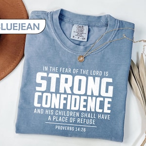 May include: A blue jean colored t-shirt with white text that reads: IN THE FEAR OF THE LORD IS STRONG CONFIDENCE AND HIS CHILDREN SHALL HAVE A PLACE OF REFUGE PROVERBS 14:26. The shirt features a crew neckline and a Comfort Colors brand label.