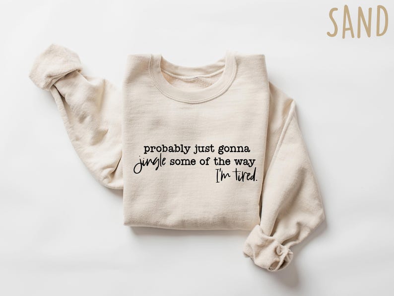 May include: Sand-colored sweatshirt with the text "probably just gonna jingle some of the way I'm tired" in black script. The long sleeves are rolled up. The sweatshirt is made of a soft material.