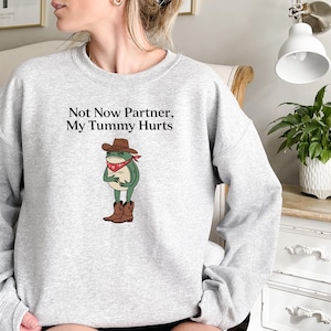 May include: Gray sweatshirt with a frog wearing a cowboy hat and bandana. The text on the sweatshirt reads "Not Now Partner, My Tummy Hurts."