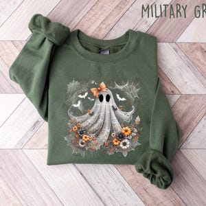 Op de afbeelding: Militairgroen sweatshirt met een grillig spookontwerp. Het spook is versierd met een strik en omgeven door bloemen, vleermuizen en spinnenwebben. De tekst "MILITARY GREEN" staat bovenaan de afbeelding.