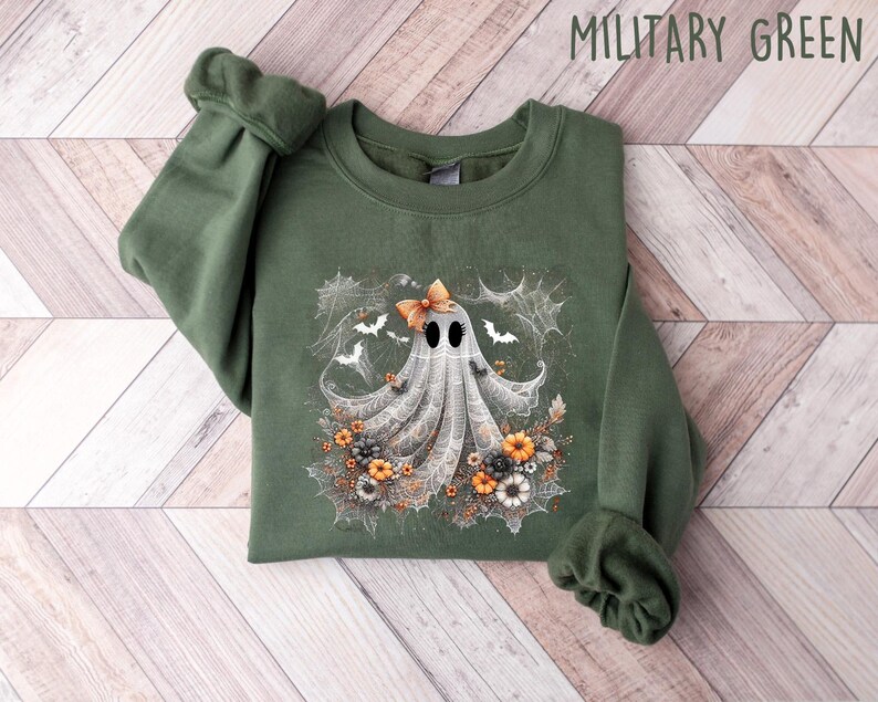 Op de afbeelding: Militairgroen sweatshirt met een grillig spookontwerp. Het spook is versierd met een strik en omgeven door bloemen, vleermuizen en spinnenwebben. De tekst "MILITARY GREEN" staat bovenaan de afbeelding.