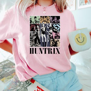 May include: Pink sweatshirt with a graphic design featuring multiple images of women in various poses, with the word "HUNTRIX" in a bold, black font. The sweatshirt is paired with light blue shorts.