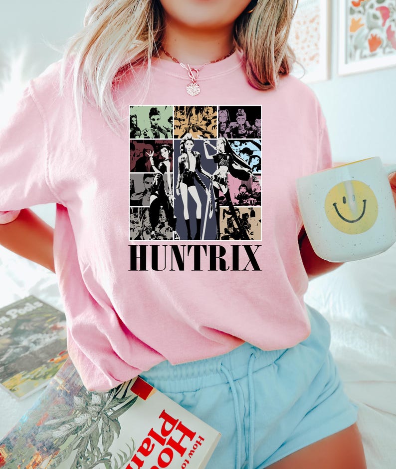 May include: Pink sweatshirt with a graphic design featuring multiple images of women in various poses, with the word "HUNTRIX" in a bold, black font. The sweatshirt is paired with light blue shorts.