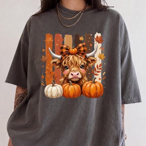 May include: A charcoal gray t-shirt featuring a graphic of a fluffy highland cow wearing a plaid bow, surrounded by orange and white pumpkins and autumn leaves against a background of vertical brushstrokes in shades of brown and orange.