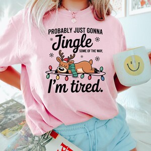 May include: A light pink sweatshirt with the text "Probably just gonna Jingle some of the way, I'm tired." A cartoon reindeer wearing a scarf and a Santa hat is lying on a string of Christmas lights. A white mug with a smiley face is held in the hand.