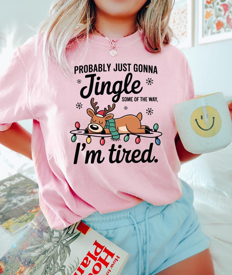 May include: A light pink sweatshirt with the text "Probably just gonna Jingle some of the way, I'm tired." A cartoon reindeer wearing a scarf and a Santa hat is lying on a string of Christmas lights. A white mug with a smiley face is held in the hand.