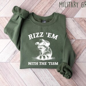 K&ouml;nnte beinhalten: Ein gr&uuml;ner Pullover mit dem Text "Rizz 'Em With The 'Tism" und einem Cartoon-Waschb&auml;ren, der einen Cowboyhut tr&auml;gt.
