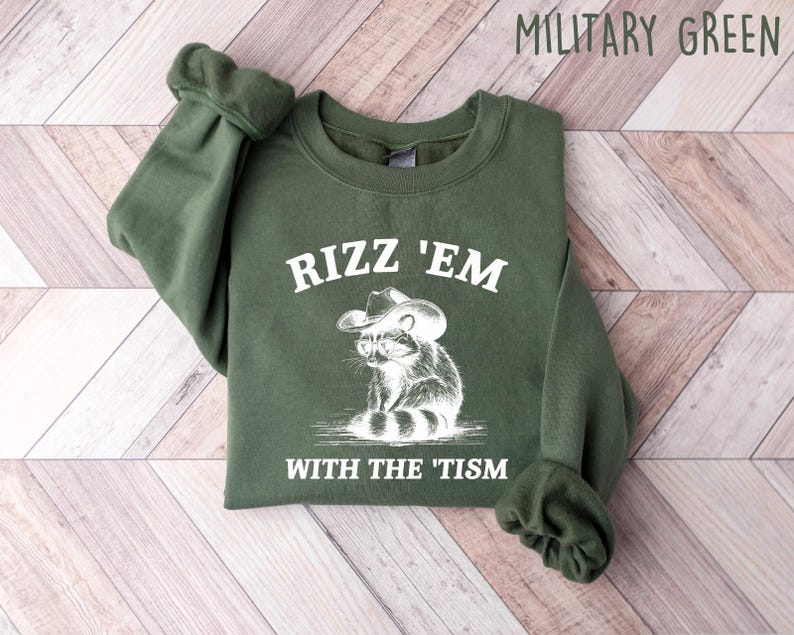 K&ouml;nnte beinhalten: Ein gr&uuml;ner Pullover mit dem Text "Rizz 'Em With The 'Tism" und einem Cartoon-Waschb&auml;ren, der einen Cowboyhut tr&auml;gt.