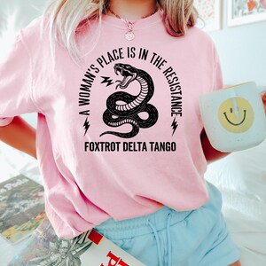 May include: A pink t-shirt with a black snake graphic and the text "A woman's place is in the resistance. Fox trot delta tango."