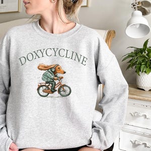 K&ouml;nnte beinhalten: Grauer Pullover mit dem Aufdruck "DOXYCYCLINE" auf der Vorderseite. Ein Cartoon-Dackel mit Kopfh&ouml;rern f&auml;hrt Fahrrad.