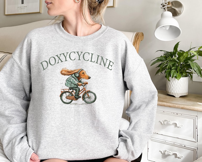 K&ouml;nnte beinhalten: Grauer Pullover mit dem Aufdruck "DOXYCYCLINE" auf der Vorderseite. Ein Cartoon-Dackel mit Kopfh&ouml;rern f&auml;hrt Fahrrad.