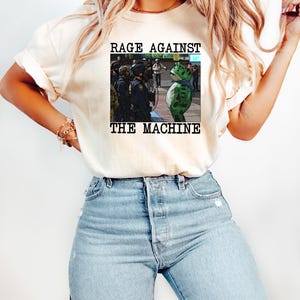 May include: Cream-colored t-shirt featuring the text "RAGE AGAINST THE MACHINE" above a graphic of a frog in front of police officers. The frog is green with a blue scarf. The shirt is paired with blue jeans.