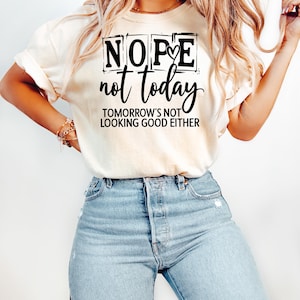 May include: Cream-colored t-shirt with the text "NOPE not today TOMORROW'S NOT LOOKING GOOD EITHER" in black script. The shirt is paired with light blue denim jeans. The overall style is casual and relaxed.