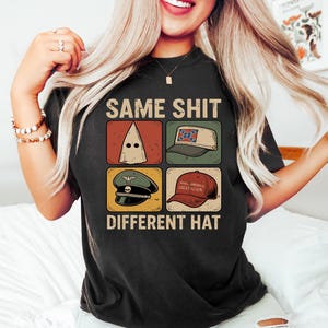 Pu&ograve; includere: T-shirt nera con la scritta "SAME SHIT DIFFERENT HAT" e quattro immagini di cappelli diversi. I cappelli includono un cappuccio bianco, un cappello con la bandiera confederata, un cappello militare e un cappello rosso "Make America Great Again".