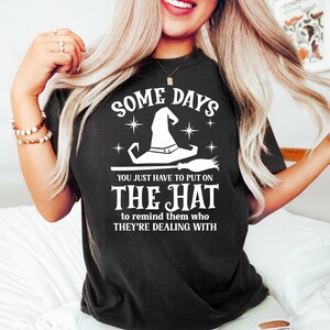 May include: Black t-shirt with the text "SOME DAYS" above a white witch hat and broom graphic. Below the graphic, the text reads "YOU JUST HAVE TO PUT ON THE HAT to remind them who THEY'RE DEALING WITH."