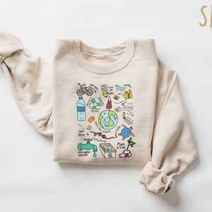 Pode incluir: Uma camisola cor de areia com um gr&aacute;fico colorido de s&iacute;mbolos de conserva&ccedil;&atilde;o ambiental. O design inclui uma bicicleta, uma garrafa de &aacute;gua, um s&iacute;mbolo de reciclagem, um globo e o texto "don't litter" e "save the planet".