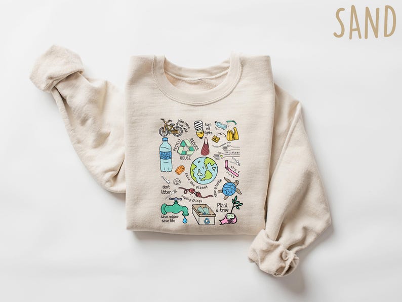 Pode incluir: Uma camisola cor de areia com um gr&aacute;fico colorido de s&iacute;mbolos de conserva&ccedil;&atilde;o ambiental. O design inclui uma bicicleta, uma garrafa de &aacute;gua, um s&iacute;mbolo de reciclagem, um globo e o texto "don't litter" e "save the planet".