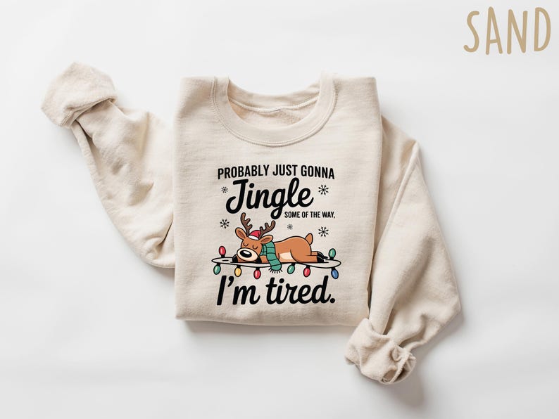May include: A sand-colored sweatshirt with the text "Probably Just Gonna Jingle Some of the Way, I'm Tired." A cartoon reindeer wearing a Santa hat and scarf is lying on a string of Christmas lights.