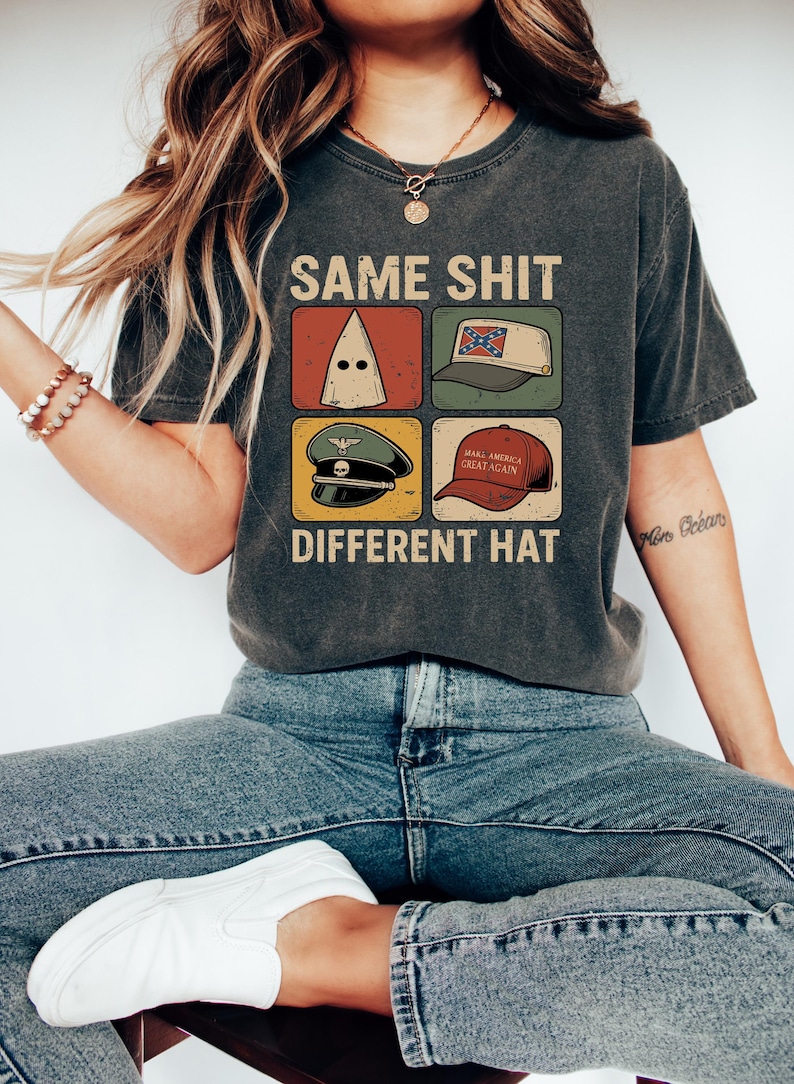 Pu&ograve; includere: T-shirt grigio scuro con la scritta "SAME SHIT DIFFERENT HAT" e quattro immagini grafiche di diversi cappelli. I cappelli includono un cappuccio bianco, un cappello con la bandiera confederata, un cappello militare e un cappello "Make America Great Again".