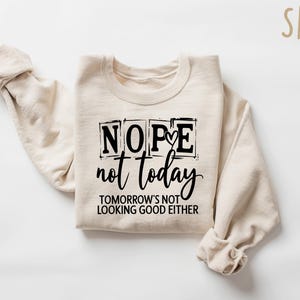 May include: Sand-colored sweatshirt with the text "Nope not today" in black, with the phrase "Tomorrow's not looking good either" below. The sweatshirt is folded with the sleeves rolled up.