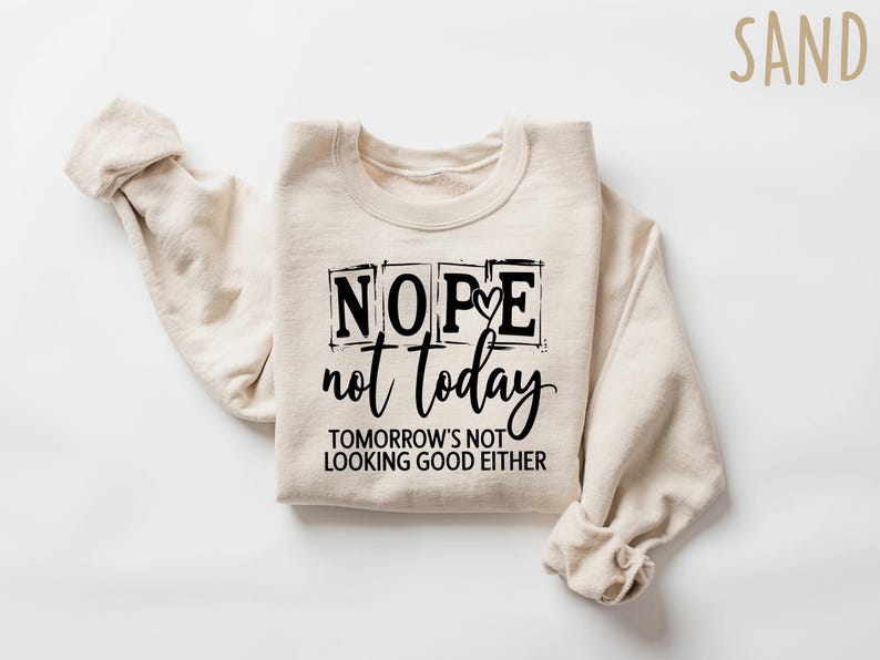 May include: Sand-colored sweatshirt with the text "Nope not today" in black, with the phrase "Tomorrow's not looking good either" below. The sweatshirt is folded with the sleeves rolled up.