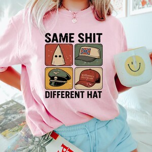 Pu&ograve; includere: Maglietta rosa con la scritta "SAME SHIT DIFFERENT HAT" e quattro immagini di cappelli. Una persona tiene in mano una tazza bianca con una faccina gialla. La persona indossa pantaloncini blu.