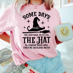 May include: A light pink sweatshirt with the text "SOME DAYS YOU JUST HAVE TO PUT ON THE HAT to remind them who THEY'RE DEALING WITH." A witch hat and broom graphic are featured. A white mug with a smiley face is held.