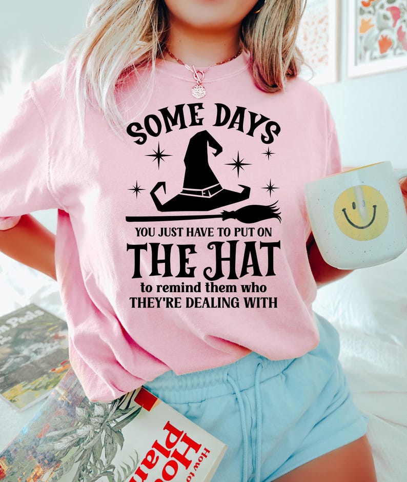 May include: A light pink sweatshirt with the text "SOME DAYS YOU JUST HAVE TO PUT ON THE HAT to remind them who THEY'RE DEALING WITH." A witch hat and broom graphic are featured. A white mug with a smiley face is held.