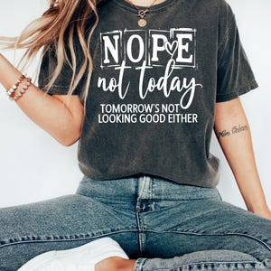 May include: Dark gray t-shirt with the text "Nope not today" in a white, bold font. The shirt also includes the phrase "Tomorrow's not looking good either". The model is wearing blue jeans and white shoes.