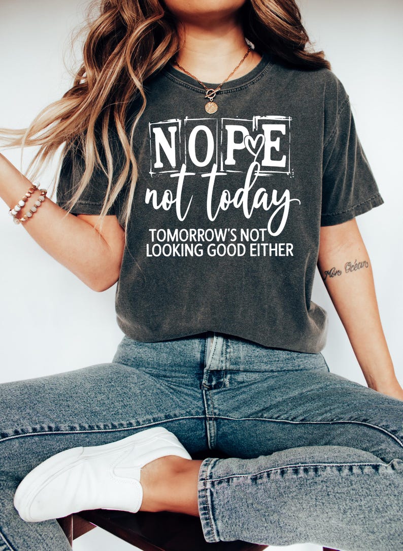 May include: Dark gray t-shirt with the text "Nope not today" in a white, bold font. The shirt also includes the phrase "Tomorrow's not looking good either". The model is wearing blue jeans and white shoes.
