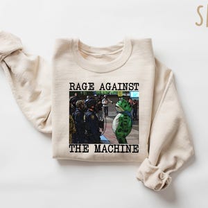 May include: Sand-colored sweatshirt with the text "RAGE AGAINST THE MACHINE" above an image of a frog-like character facing police. The frog is green with a blue scarf. The sweatshirt has long sleeves.