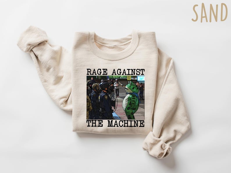 May include: Sand-colored sweatshirt with the text "RAGE AGAINST THE MACHINE" above an image of a frog-like character facing police. The frog is green with a blue scarf. The sweatshirt has long sleeves.