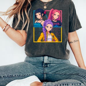 May include: Dark gray t-shirt featuring a colorful anime-style graphic of three women. The graphic includes the word "VENOM" and is framed by yellow, blue, and pink borders. The shirt is worn with blue jeans and white sneakers.