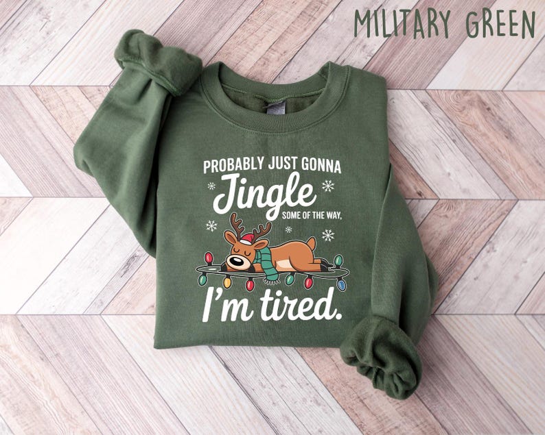 May include: Military green sweatshirt with the text "Probably just gonna Jingle some of the way, I'm tired." A cartoon reindeer wearing a scarf and a Santa hat is lying on a string of Christmas lights. The sweatshirt is folded on a light wood surface.