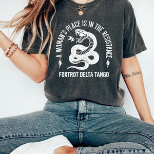 May include: A dark gray t-shirt with a white graphic of a snake with the text "A woman's place is in the resistance. Fox Trot Delta Tango".