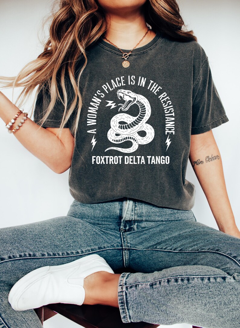 May include: A dark gray t-shirt with a white graphic of a snake with the text "A woman's place is in the resistance. Fox Trot Delta Tango".