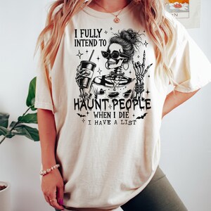 May include: Cream-colored t-shirt with a black graphic of a skeleton wearing sunglasses and holding a drink. The text reads "I FULLY INTEND TO HAUNT PEOPLE WHEN I DIE I HAVE A LIST."