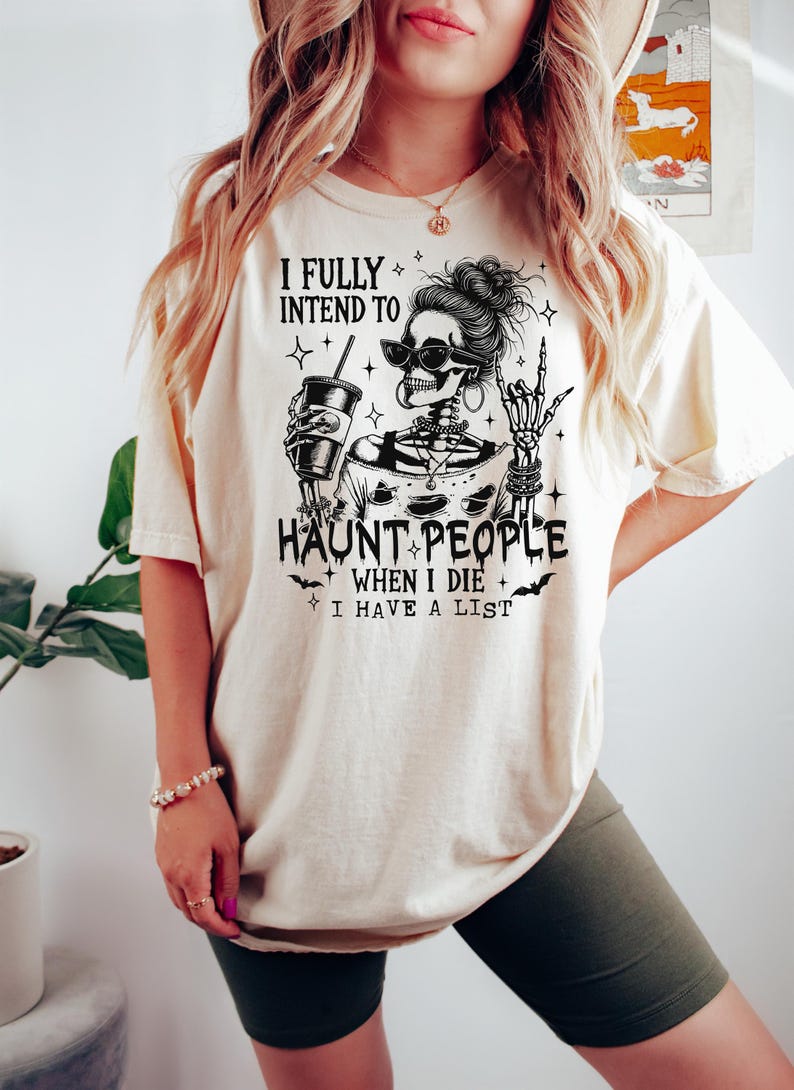May include: Cream-colored t-shirt with a black graphic of a skeleton wearing sunglasses and holding a drink. The text reads "I FULLY INTEND TO HAUNT PEOPLE WHEN I DIE I HAVE A LIST."