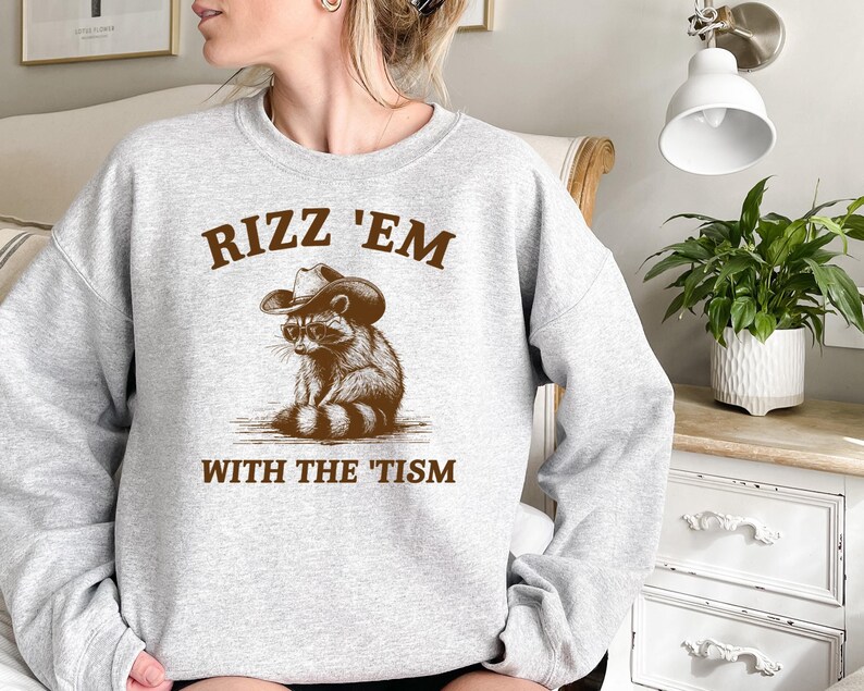 K&ouml;nnte beinhalten: Grauer Pullover mit einem braunen Waschb&auml;ren, der einen Cowboyhut und eine Sonnenbrille tr&auml;gt. Der Text "Rizz 'Em With The 'Tism" ist auf dem Pullover aufgedruckt.